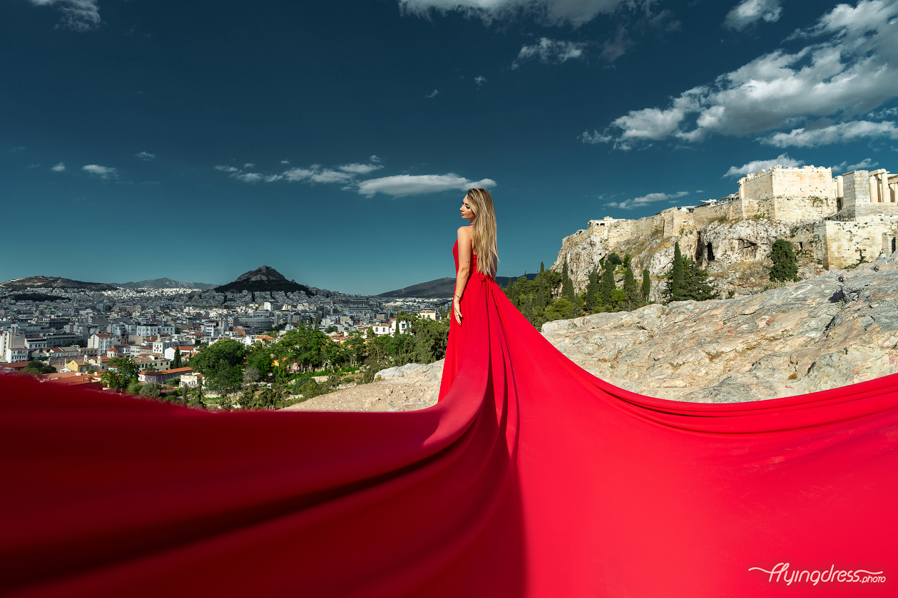 A woman in a flowing red dress poses with the cityscape of Athens and the ancient ruins of the Acropolis in the background, under a partly cloudy sky.